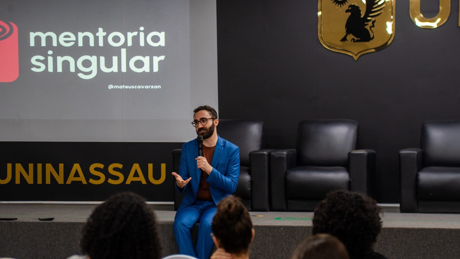 Mateus Cavarzan sentado na beira do palco da UNINASSAU, falando com a plateia