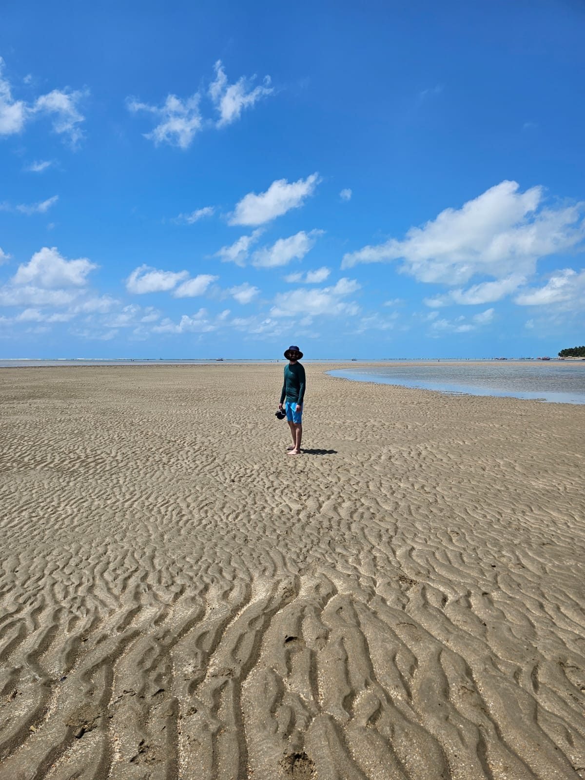 Mateus Cavarzan de pé na areia da Praia do Patacho, em Alagoas, na maré baixa, sob céu azul com nuvens
