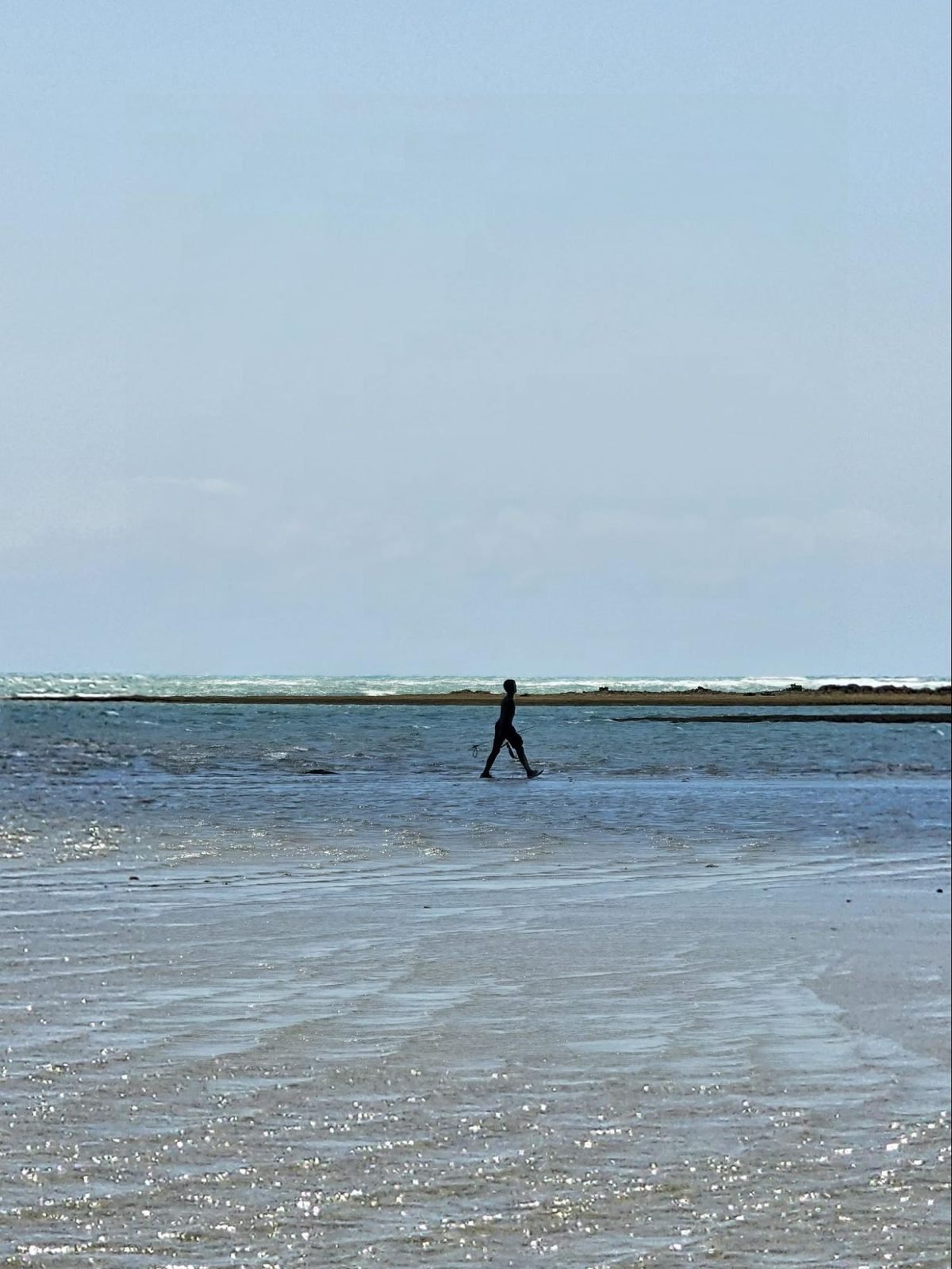 Pessoa caminhando com a água nos tornozelos na maré baixa da Praia do Patacho, com o horizonte ao fundo
